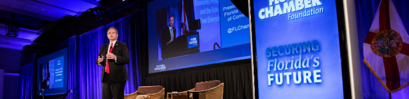 Gentleman on stage with blue curtains behind him and screen stating title of  meeting. 