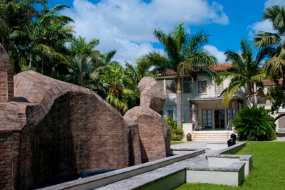 Photo of a White Spanish style house with brown roofing Palm trees and a large red brick sculpture 