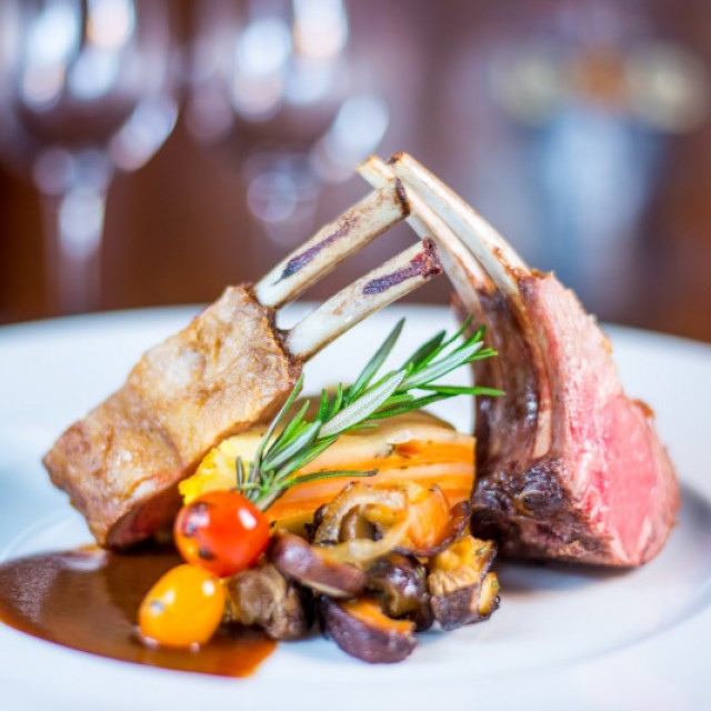 Photo of a plated entrée of 4 baby lamb chops standing up on a plate with a potato casserole in the middle and onion mushroom and grape tomato side.